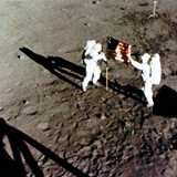 Neil Armstrong and Buzz Aldrin by the flag. A frame from the 16 mm film taken out the LM window. TV in the back